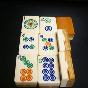Op de afbeelding: Een verzameling ivoorkleurige Mahjong-tegels met kleurrijke cirkelvormige ontwerpen en cijfers. De tegels zijn in een stapel gerangschikt en tonen de ingewikkelde details en levendige kleuren van de speelstukken. De set bevat een houten doos.