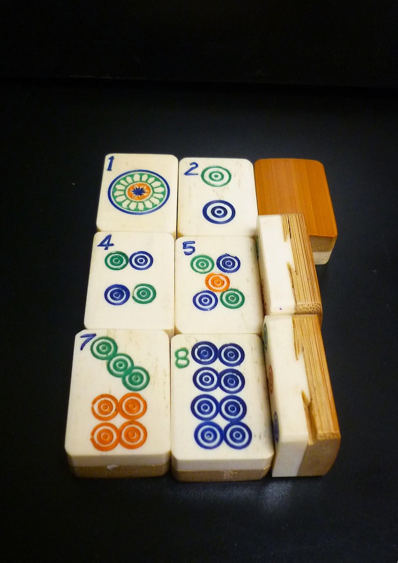 Op de afbeelding: Een verzameling ivoorkleurige Mahjong-tegels met kleurrijke cirkelvormige ontwerpen en cijfers. De tegels zijn in een stapel gerangschikt en tonen de ingewikkelde details en levendige kleuren van de speelstukken. De set bevat een houten doos.