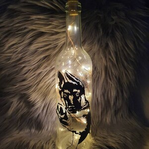 May include: A clear glass bottle with a cork top and a silhouette of a German Shepherd puppy inside. The bottle is filled with warm white fairy lights.