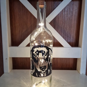 May include: A clear glass wine bottle with a brown cork stopper, illuminated by string lights. A black and white dachshund dog graphic is on the bottle. The bottle is displayed on a white chair.