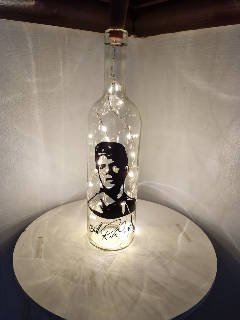 May include: A clear glass bottle with a black silhouette of a person's face and a signature. The bottle is filled with white fairy lights.