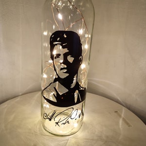 May include: A clear glass bottle with a black silhouette of a man's face and signature. The bottle is lit with warm white fairy lights.