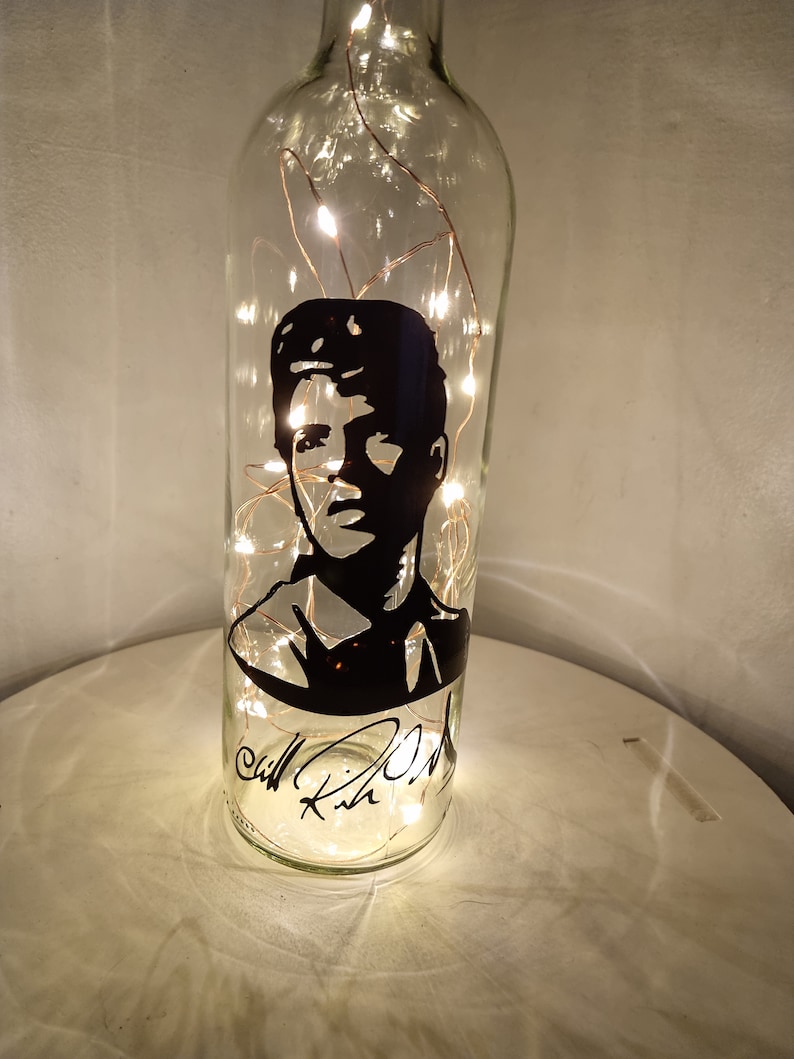 May include: A clear glass bottle with a black silhouette of a man's face and signature. The bottle is lit with warm white fairy lights.