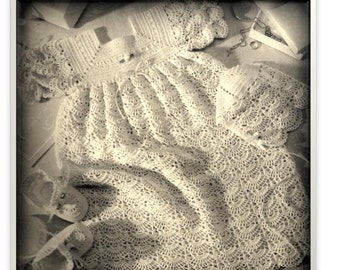 May include: A white crocheted baby gown, bonnet, and booties. The gown has a scalloped edge and is laid out on a surface with a jewelry box and chain.