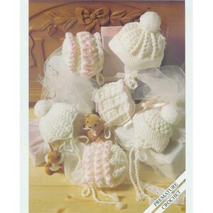 May include: A crochet pattern for a baby hat, with a white hat with a pom pom on top, and a pink and white hat with a ruffle. The pattern is called "Snuggly Double Knitting" and is for premature to 6 months. The pattern is from Sirdar.