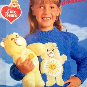 May include: A young girl wearing a blue knitted sweater with a Care Bear design. The sweater features a yellow Care Bear with a smiling sun on its belly. The girl is holding a yellow Care Bear plush toy.