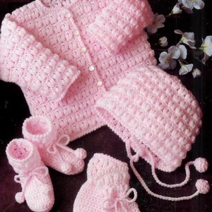 May include: A pink crocheted baby set comprising a cardigan, hat, booties, and mittens. The set is made with a textured stitch and features white buttons on the cardigan.