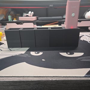 May include: A matte black rectangular organizer with four square compartments, one of which holds a light pink electric hair clipper with a silver blade and a small circular button, featuring the word CLIPPER printed on the front of the device.
