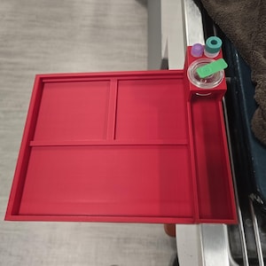 May include: A red plastic organizer tray featuring three main compartments and a side section with a circular holder. The circular holder contains a clear plastic lid with a green tab, a small purple cap, and a small teal cap.