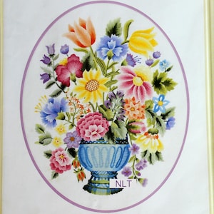 May include: A framed embroidery piece featuring a vibrant bouquet of multi-colored flowers in a blue vase. The floral arrangement includes shades of orange, yellow, pink, purple, and blue, set within a purple oval border. The text NLT is visible.