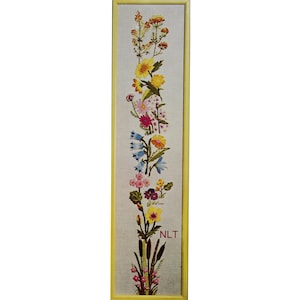 May include: A framed floral embroidery featuring a vertical arrangement of colorful flowers. The embroidery includes yellow, pink, blue, and purple blooms, with green leaves and stems. The frame is a light yellow color, and the text "NLT" is visible.