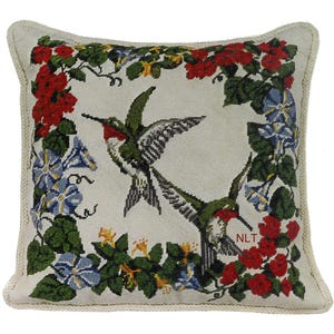 May include: Square decorative pillow with a needlepoint design featuring two hummingbirds in flight, surrounded by a floral border of red, blue, and yellow flowers and green leaves. The pillow has a cream-colored background and a knitted border.