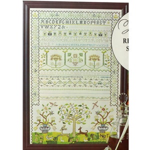 May include: Framed cross-stitch sampler with a dark brown wooden frame. The sampler features an alphabet, floral and tree motifs, and the text "ABCDEFGHIKLMNOPARST VWXYZ&." The color palette includes green, beige, and brown.