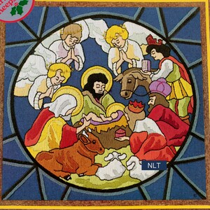 May include: A circular embroidery piece depicting a nativity scene with figures in colorful robes, angels, a donkey, a cow, and a sheep. The design is framed by dark blue lines resembling stained glass, with the text NLT in a blue box.