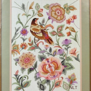 May include: A framed embroidery piece on a light fabric background featuring a brown and orange bird perched among various colorful flowers in shades of pink, orange, purple, and green. The letters NLT are stitched in the bottom right corner.
