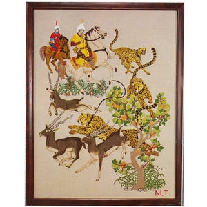 May include: A framed embroidery depicting a hunting scene with two riders on horseback, one in red and one in yellow, pursuing several cheetahs and antelopes near a tree. The scene is set on a light beige fabric background with the letters NLT.