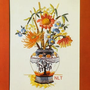 May include: An embroidery piece on off-white fabric featuring a vase with a black tree silhouette and orange floral patterns. The vase holds a bouquet of orange, yellow, and blue flowers with green stems. The letters NLT are stitched in red.