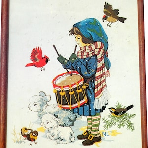 May include: A framed embroidery depicting a child in a blue coat, striped scarf, and green gloves playing a drum. The scene includes two white lambs, two small birds on the ground, a red cardinal, a flying bird, and the letters NLT in the corner.