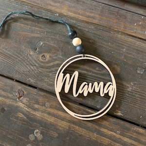 May include: A wooden circle ornament with the word "Mama" written in cursive. The ornament is hanging from a black string with black and beige beads.