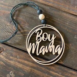 May include: A wooden circle ornament with the text "Boy Mama" in a black script font. The ornament is hanging from a black string with black and white beads.