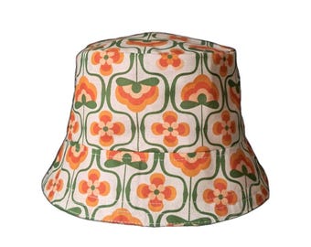 May include: A white bucket hat with a repeating floral pattern in orange and green. The hat has a rounded crown and a downward-sloping brim. The floral design is reminiscent of the 1970s.