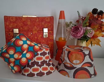 May include: Three bucket hats featuring retro-inspired geometric patterns in shades of orange, brown, cream, and teal. The hats are displayed alongside a rectangular orange patterned case, an orange lava lamp, and a bouquet of artificial flowers.