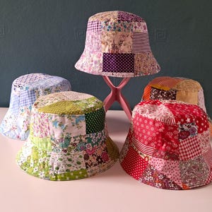 May include: Four bucket hats featuring patchwork designs made from various fabric squares. The hats display diverse patterns including floral, polka dot, gingham, and striped prints in shades of pink, red, green, blue, and white. One hat is elevated on a pink stand.
