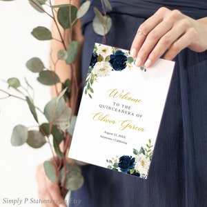 May include: A white card with gold lettering announcing a Quinceañera for Olivia Garcia on August 25, 20XX in Anytown, California. The card is decorated with blue and white flowers.
