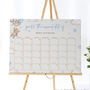 Op de afbeelding: Een lichtblauw en bruin baby shower-spelbord met een kalender waarop gasten de aankomstdatum van Baby Stainton kunnen raden. Het bord toont een cartoon teddybeer met blauwe ballonnen en sterren.
