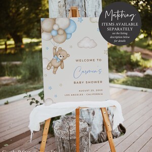 Op de afbeelding: Een welkomstbord voor een babyshower met een blauwe en bruine teddybeer die ballonnen vasthoudt. Het bord staat "Welkom op Carmen's babyshower" met de datum en locatie eronder.