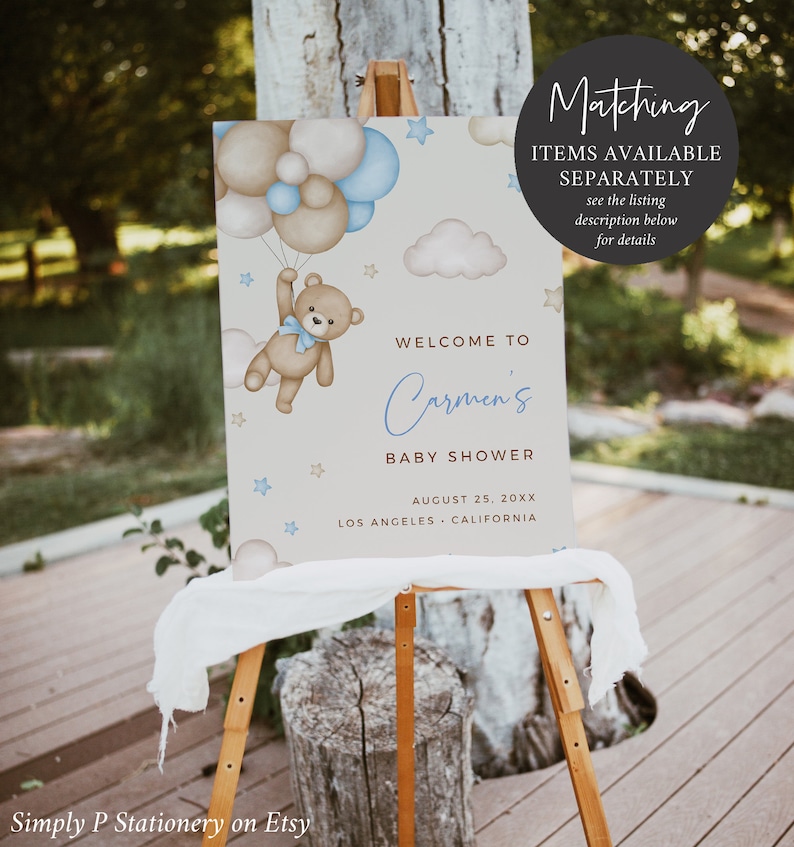 Op de afbeelding: Een welkomstbord voor een babyshower met een blauwe en bruine teddybeer die ballonnen vasthoudt. Het bord staat "Welkom op Carmen's babyshower" met de datum en locatie eronder.