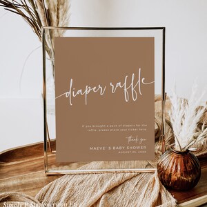 May include: A brown sign with white text that reads "diaper raffle". The sign is in a gold frame and is sitting on a wooden table. The text below the title reads "If you brought a pack of diapers for the raffle, please place your ticket here. Thank you. MAEVE'S BABY SHOWER AUGUST 25, 20XX"