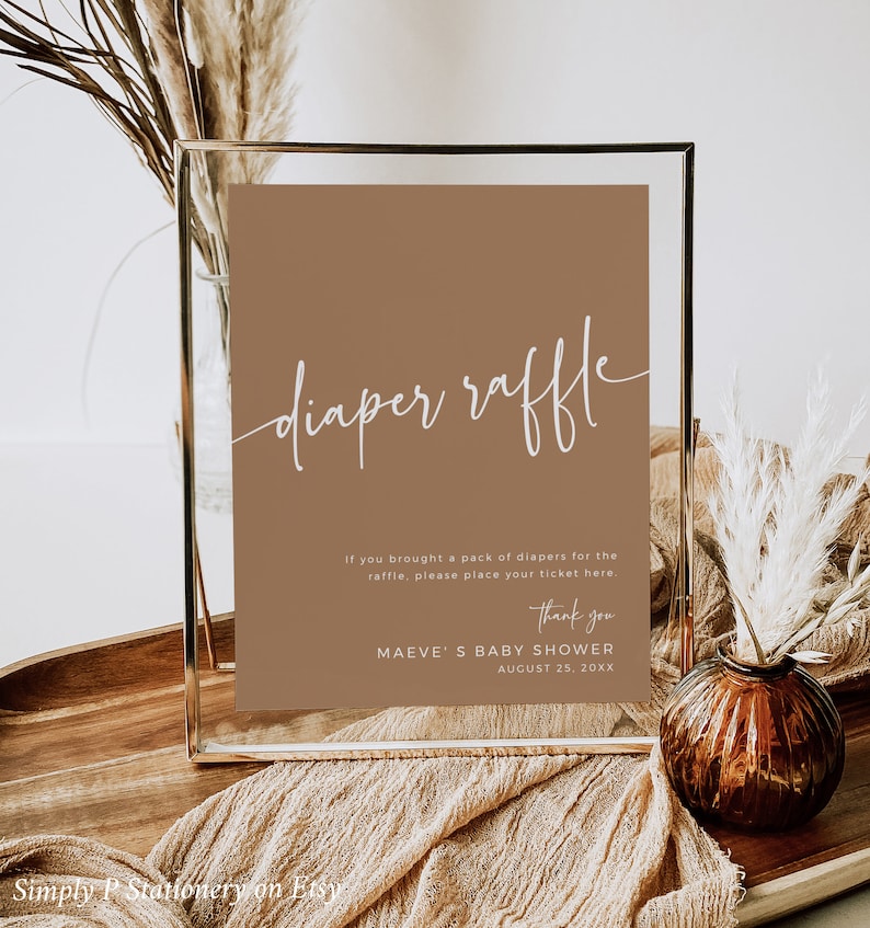 May include: A brown sign with white text that reads "diaper raffle". The sign is in a gold frame and is sitting on a wooden table. The text below the title reads "If you brought a pack of diapers for the raffle, please place your ticket here. Thank you. MAEVE'S BABY SHOWER AUGUST 25, 20XX"