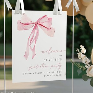 May include: A white sign with a pink watercolor bow on top. The sign says "Welcome to Blythe's Graduation Party Cedar Valley High School Class of 20XX".
