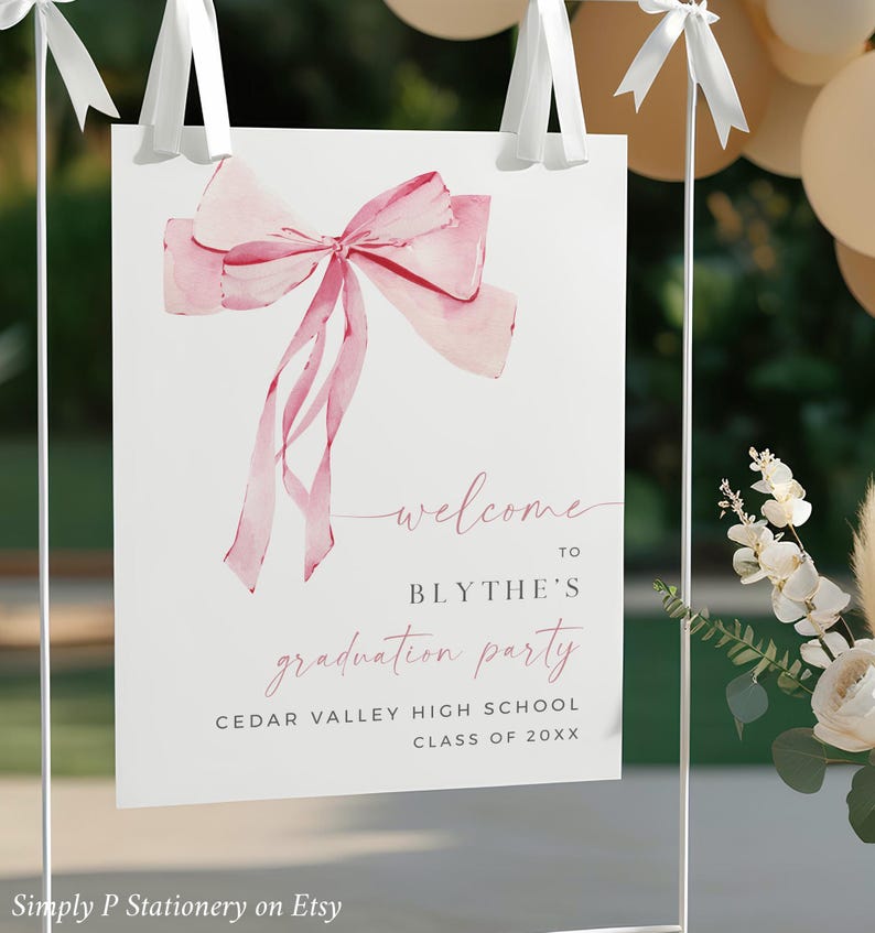 May include: A white sign with a pink watercolor bow on top. The sign says "Welcome to Blythe's Graduation Party Cedar Valley High School Class of 20XX".