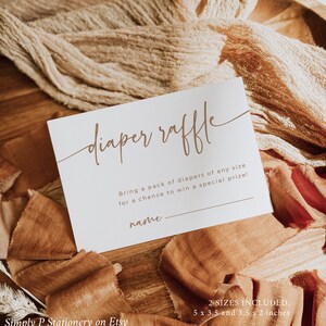 May include: A white card with brown text that says "diaper raffle". The card has a line for a name and the text "Bring a pack of diapers of any size for a chance to win a special prize!"  The card is on a wooden surface with brown fabric.