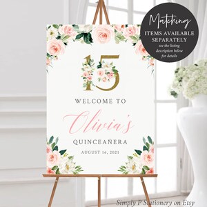 May include: A white welcome sign with pink and white flowers and greenery. The sign has gold glitter lettering that says "15" and "Welcome to Olivia's Quincea&ntilde;era August 16, 2021".