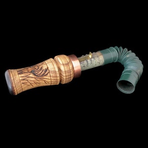 May include: A wooden duck call with a green and brown telescoping extension. The call has a carved design of a duck on the wooden portion.