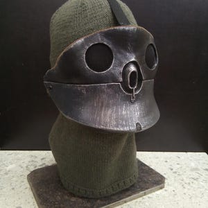 Może przedstawiać: Ciemnooliwkowa czapka z dzianiny noszona z maską na twarz o metalicznym wyglądzie. Maska jest w kolorze ciemnoszarym z wyblakłym, szczotkowanym wykończeniem, posiada dwa okrągłe otwory na oczy pokryte czarną siatką i centralny otwór na nos.