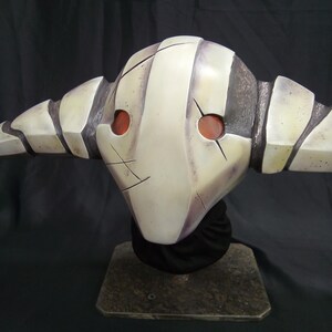 May include: A white and gray mask with red eyes and a striped design. The mask is on a black stand.
