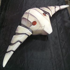 May include: A white and gray mask with a pointed, curved shape and two red circles. The mask has a textured surface and appears to be made of a hard material.