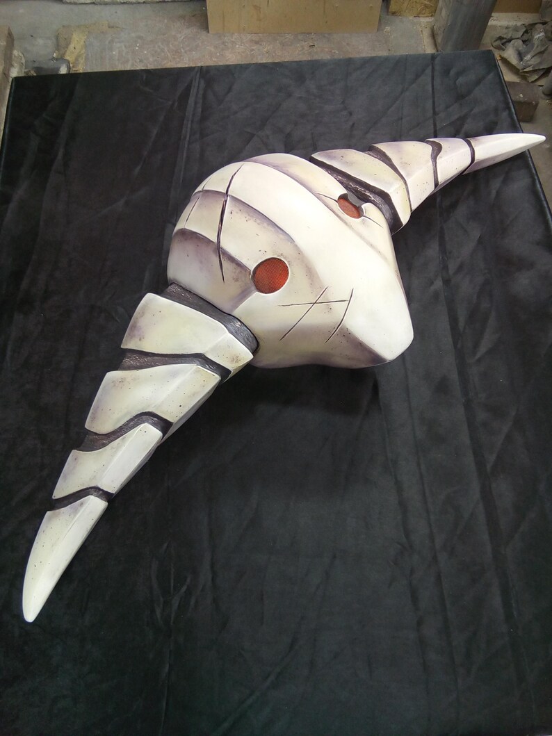 May include: A white and gray mask with a pointed, curved shape and two red circles. The mask has a textured surface and appears to be made of a hard material.