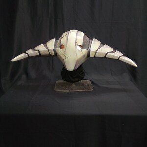 May include: A white and gray mask with horns, resembling a creature with a skeletal face. The mask is on a black stand against a black background.
