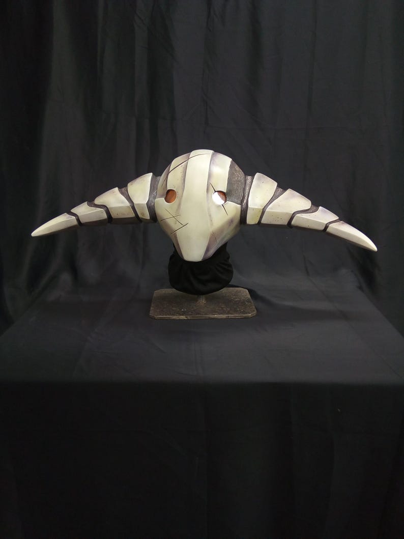 May include: A white and gray mask with horns, resembling a creature with a skeletal face. The mask is on a black stand against a black background.