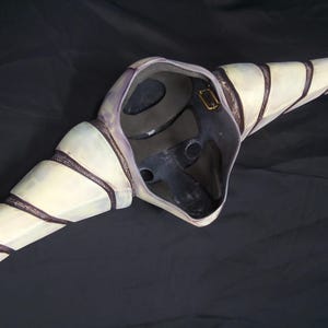 May include: A white and purple mask with black stripes and a textured surface. The mask has a large beak and a hole for the eyes.