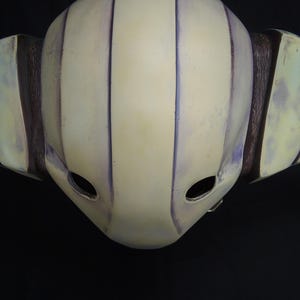 May include: A white helmet with purple stripes and two eye holes. The helmet is made of a hard material and has a smooth surface.