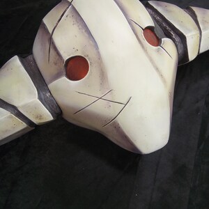 May include: A white, textured, helmet-like mask with two red, circular eye holes. The mask has a jagged, angular design and appears to be made of a hard material.