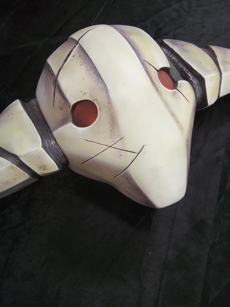 May include: A white, textured, helmet-like mask with two red, circular eye holes. The mask has a jagged, angular design and appears to be made of a hard material.