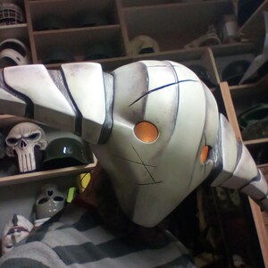 May include: A white and gray mask with a long, curved horn. The mask has two orange eyes and a black design on the front. The mask is being held by a person.