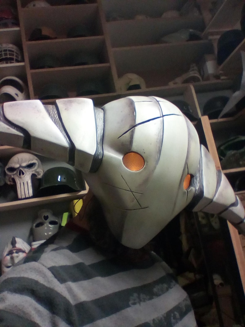 May include: A white and gray mask with a long, curved horn. The mask has two orange eyes and a black design on the front. The mask is being held by a person.